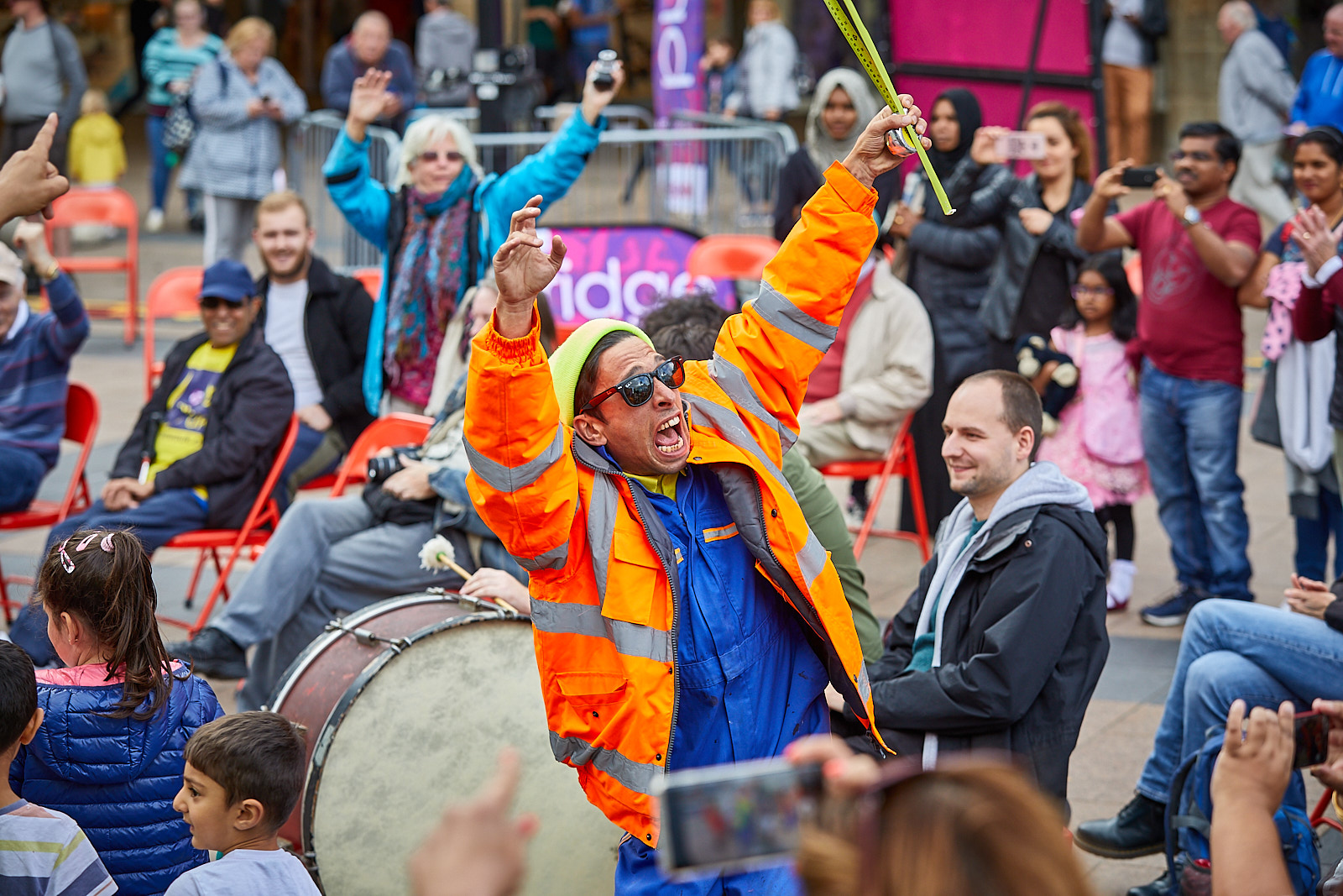 An actor dressed in high-vis jacket and overalls raises his hands to the crowd whilst pulling a funny expression. Behind him a lady copies his action. They are surrounded by musicians and audiences. People are smiling.
