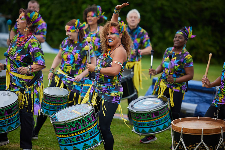 A group of drummers wear bright, African-inspired costume and face paints. A woman with feathers in her hair raises her hands to the sky.
