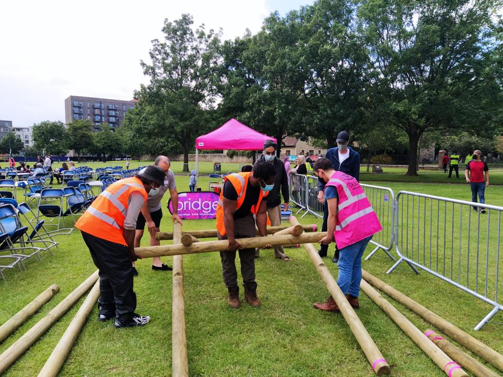 In an urban park, a group of men attempt to put together a self-supporting bridge made of wooden poles. They are wearing high-vis jackets and face masks.