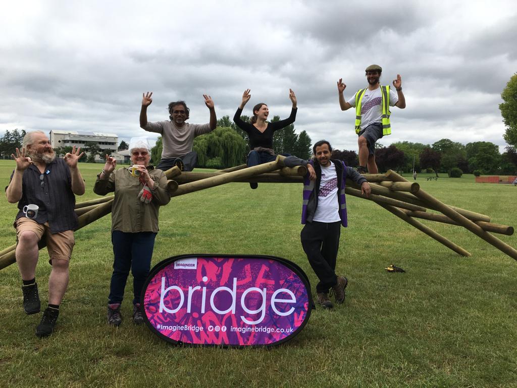6 people are sitting on or around a self-supporting bridge made of wooden poles. They have their hands in the air in celebration. The bridge is holding their weight despite not having any glue or nails to hold it together!